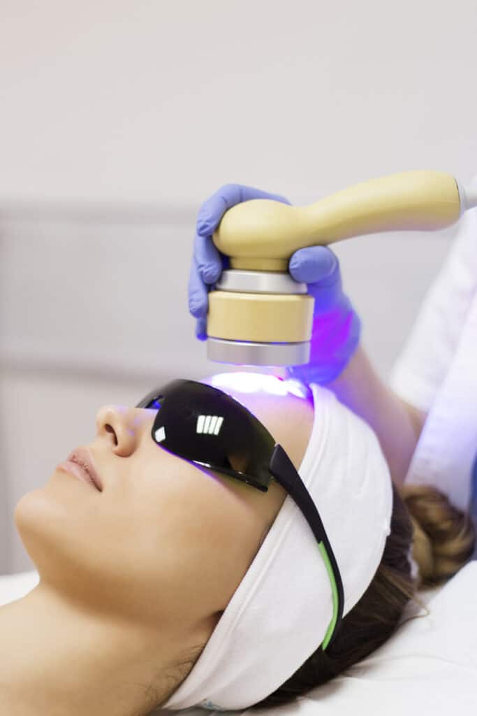 A woman at a dermatological clinic undergoing ultraviolet light therapy on her face, she is wearing protective eye wear. 