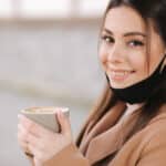 Beautiful woman takes off protective mask for drinking coffee. Female sitting outdoors on terrace and drink coffee. Quarantine, self distancing.