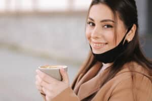 Beautiful woman takes off protective mask for drinking coffee. Female sitting outdoors on terrace and drink coffee. Quarantine, self distancing.