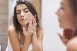 young woman with acne problem looking in mirror 