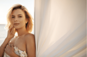 Short-haired blonde looks tenderly at camera, dressed in white sundress with bare shoulders.